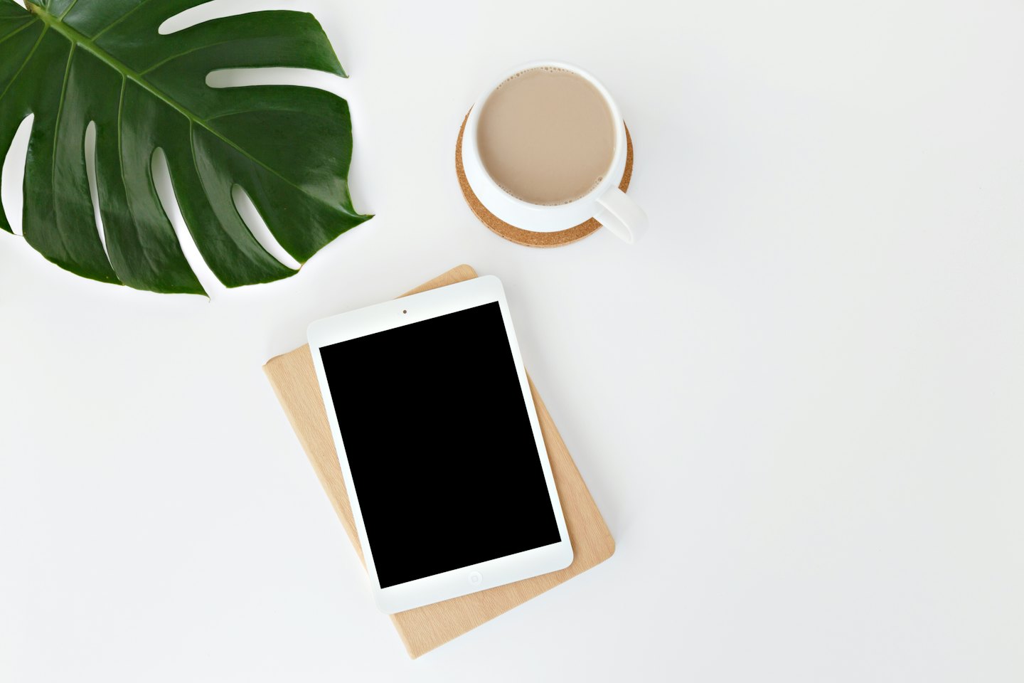 Minimalistischer Schreibtisch mit Tablet, Kaffee und Monstera-Blatt.
